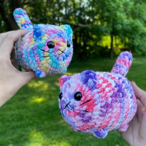 May include: Two round, plush, crocheted cat figures made from variegated yarn. One cat features a mix of blue, yellow, and pink tones, while the other displays shades of purple, pink, and white. Both have black plastic eyes and stitched whiskers.