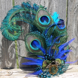 May include: A decorative headpiece featuring peacock feathers in shades of blue and green. The design includes large peacock eye feathers, smaller blue feathers, and a central embellishment with a gold-toned frame and a green stone.