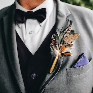 May include: A grey suit jacket with a black bow tie, a black waistcoat, and a purple pocket square. A boutonniere made of feathers and a gold bullet casing is pinned to the lapel.