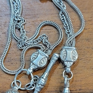 Könnte beinhalten: Eine silberfarbene Uhrenkette mit mehreren Strängen und dekorativen Elementen. Die Kette hat einen detaillierten Verschluss, einen kleinen Ball und ein zylindrisches Teil. Das aufwendige Design deutet auf einen Vintage- oder Antik-Stil hin.