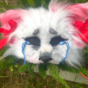 May include: A white and pink faux fur animal mask featuring large black eye sockets, a black nose, and two blue sculpted teardrops beneath the eyes. The mask is adorned with two bright red ribbons tied on each side and small grey horns.