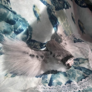 May include: A plush, gray and white seal head with a pink nose and black whiskers rests on a blue and white patterned blanket. The blanket features a sea turtle and fish design. White lace trim is visible.