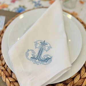 May include: White linen napkin with a blue embroidered monogram of the letter "L" in a decorative font. The napkin is folded and placed on a white plate, which is on top of a woven brown placemat.