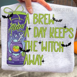 May include: A light gray t-shirt featuring a graphic of a purple Alani Witch's Brew energy drink can with a green bow, bats, and stars. The text reads, A BREW A DAY KEEPS THE WITCH AWAY, in green lettering. The design includes a cauldron and frog.