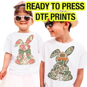 May include: Two children wearing white t-shirts with bunny designs. One bunny is floral with a green bow, the other is camouflage with a hat that says "Egg Minor". The text "READY TO PRESS DTF PRINTS" is at the top.