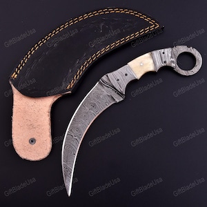 May include: A curved karambit knife with a Damascus steel blade and a bone handle, accompanied by a black leather sheath. The blade has a distinctive swirling pattern. The sheath is black with a tan leather belt loop.