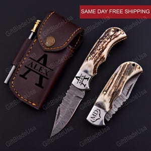 May include: A personalized pocket knife set with a leather sheath. The set includes two knives with patterned blades and antler handles. The sheath is brown with the name "ALEX" engraved. A small metal tool is also included.