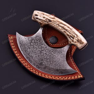 May include: A Damascus steel cleaver knife with a curved blade and a handle made of light-colored antler. The knife rests in a brown leather sheath with a stitched border. The blade has a unique pattern.