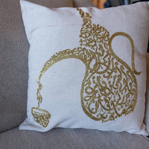 May include: A square, off-white decorative pillow featuring a gold-colored design of a traditional coffee pot and cup. The design is made up of intricate patterns and calligraphy. The pillow is placed on a neutral-colored sofa.