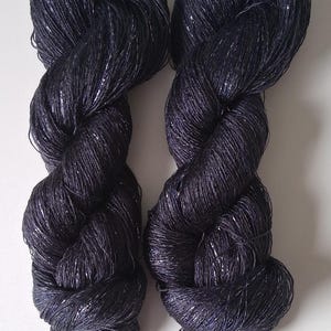 May include: Two skeins of dark navy blue yarn with a subtle metallic silver thread woven throughout. The yarn is twisted into a rope-like shape, and the texture of the yarn is visible. The background is plain white.