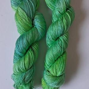May include: Two skeins of green yarn with a gradient of colours from light to dark. The yarn is twisted into a rope-like shape, with a mix of light green, dark green, and a hint of blue.