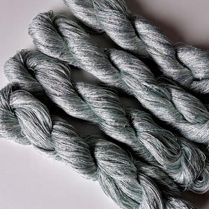 May include: Close-up of several skeins of silver-gray yarn. The yarn is tightly twisted, with a slight sheen and a subtle sparkle. The texture appears soft and pliable, suggesting it is suitable for various crafting projects.