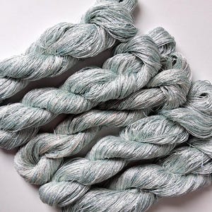 May include: Several skeins of light blue and silver yarn are arranged on a white surface. The yarn appears to be a soft, textured material, possibly silk or a similar fiber. The skeins are twisted and coiled.