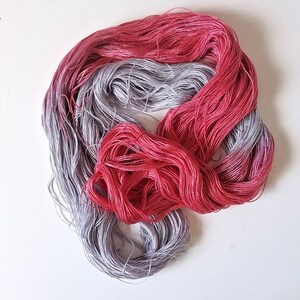 Puede incluir: Primer plano de dos madejas de hilo de bordar. Una es un degradado de rosa a rojo, la otra de gris a morado. El hilo est&aacute; dispuesto sobre una superficie blanca.