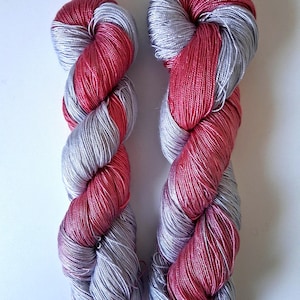 Puede incluir: Dos madejas de hilo en un patr&oacute;n retorcido. El hilo es una combinaci&oacute;n de plata y un color rosa-rojo vibrante. Las madejas se muestran sobre un fondo blanco liso, resaltando los colores y la textura del hilo.