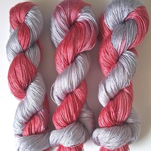 Puede incluir: Tres madejas de hilo en una configuraci&oacute;n retorcida. El hilo es una combinaci&oacute;n de plata y un vibrante color rojo-rosa. Las madejas est&aacute;n dispuestas una al lado de la otra sobre un fondo blanco, mostrando el color y la textura del hilo.