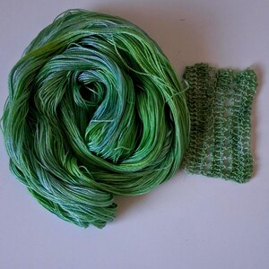 May include: A skein of green and blue variegated yarn next to a small swatch of knitted fabric. The yarn is tightly wound, displaying shades of green and blue. The knitted swatch is a lighter green with a textured, open weave.