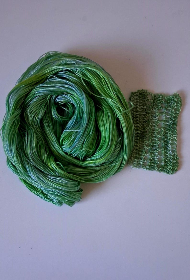 May include: A skein of green and blue variegated yarn next to a small swatch of knitted fabric. The yarn is tightly wound, displaying shades of green and blue. The knitted swatch is a lighter green with a textured, open weave.