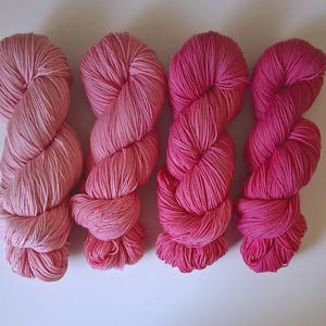 Op de afbeelding: Vier strengen garen op een rij, die een verloop van roze tinten tonen. De kleuren variëren van een bleek, zacht poederroze aan de uiterste linkerzijde tot een diep, levendig magentarood aan de rechterkant, wat een vloeiende kleurovergang vormt.