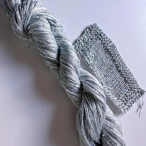 May include: A skein of silver and white yarn with a braided appearance, next to a small knitted swatch. The yarn has a metallic sheen, and the knitted piece shows the texture of the material. Ideal for knitting and crochet projects.