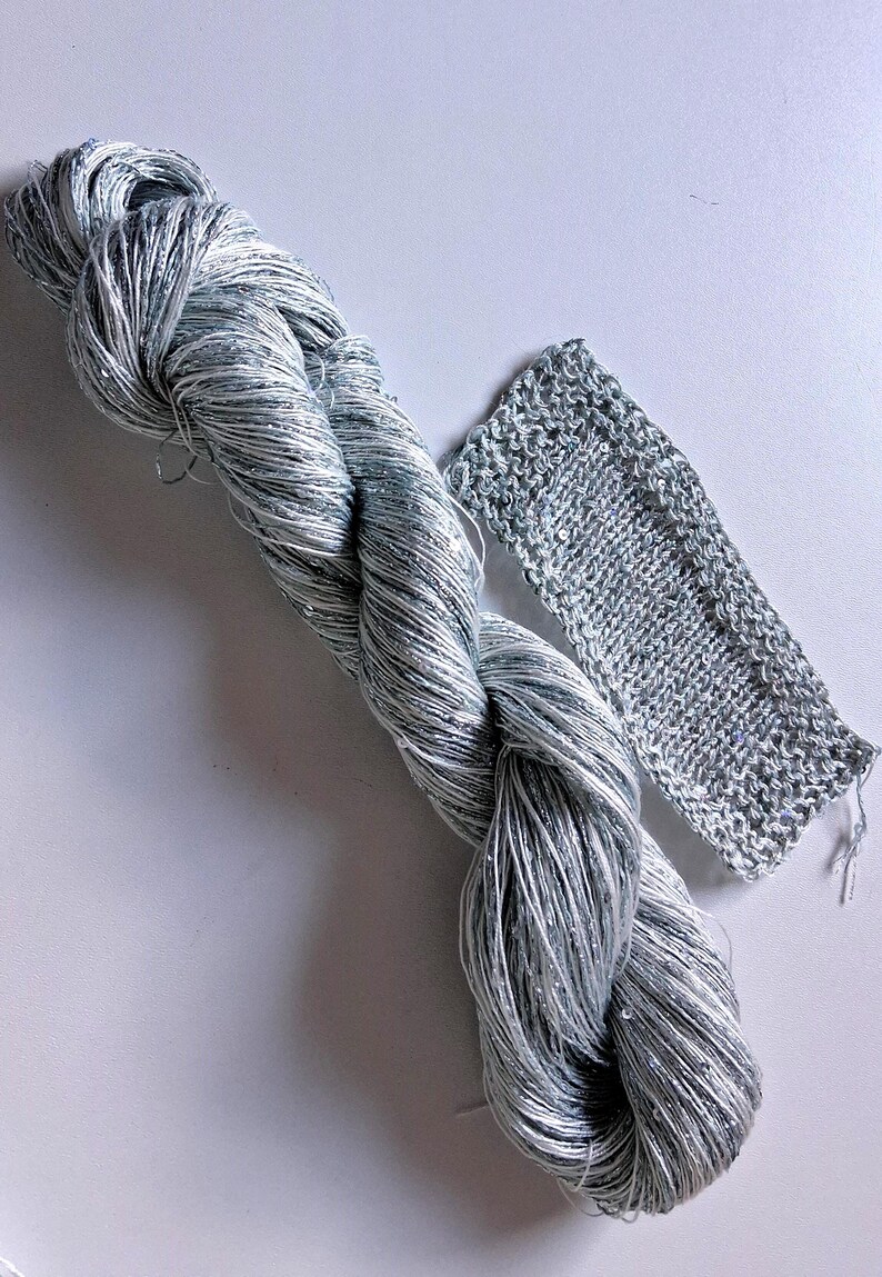 May include: A skein of silver and white yarn with a braided appearance, next to a small knitted swatch. The yarn has a metallic sheen, and the knitted piece shows the texture of the material. Ideal for knitting and crochet projects.