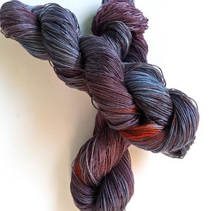 May include: A skein of hand-dyed yarn in shades of purple, blue, and orange. The yarn is twisted into a figure-eight shape, showcasing the color variations. The yarn appears soft and luxurious, suitable for knitting or crochet projects.