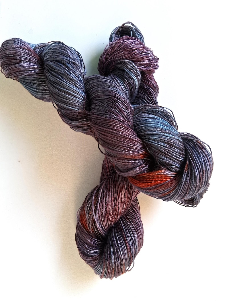 May include: A skein of hand-dyed yarn in shades of purple, blue, and orange. The yarn is twisted into a figure-eight shape, showcasing the color variations. The yarn appears soft and luxurious, suitable for knitting or crochet projects.