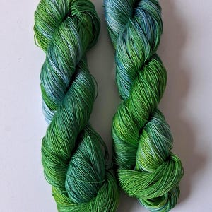 May include: Two skeins of vibrant green yarn with subtle blue and white highlights. The yarn is tightly wound, showcasing a gradient of shades from light to dark green. Ideal for knitting or crochet projects.