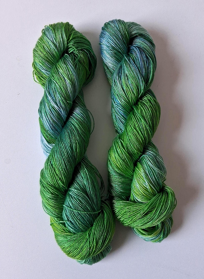 May include: Two skeins of vibrant green yarn with subtle blue and white highlights. The yarn is tightly wound, showcasing a gradient of shades from light to dark green. Ideal for knitting or crochet projects.