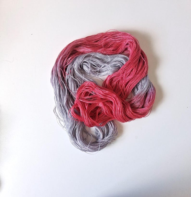 Puede incluir: Primer plano de dos madejas de hilo de bordar. Una es un degradado de rosa a rojo, la otra de gris a morado. El hilo est&aacute; dispuesto sobre una superficie blanca.