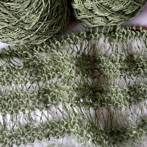 May include: Two balls of sage green yarn positioned above a piece of partially knitted fabric. The knitting features an open, airy, lace-like stitch pattern created with the same sage green yarn, showing visible loops and a delicate, sheer texture.
