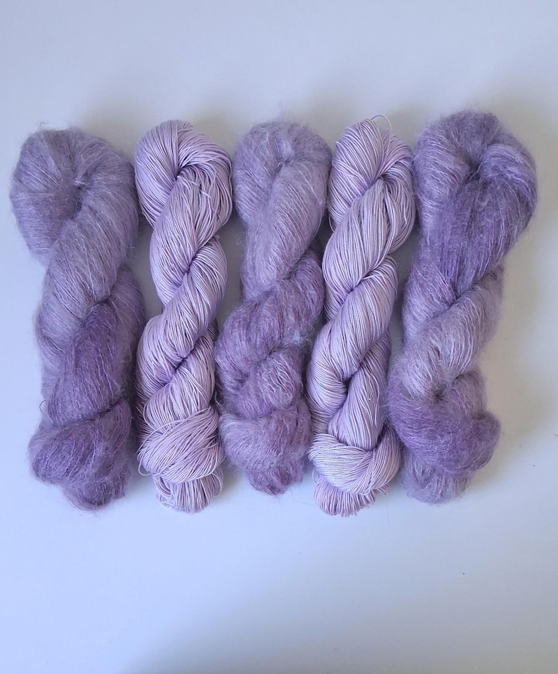 Puede incluir: Cinco madejas de hilo retorcidas dispuestas en fila, cada una con una textura suave y difusa y tonos variados de morado claro y lavanda. El hilo presenta una calidad delicada y aireada con fibras visibles en cada paquete individual.