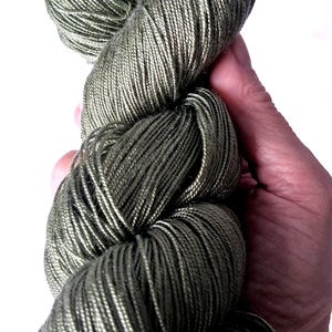 May include: A hand holds a skein of olive green yarn. The yarn is tightly wound, revealing individual strands with a subtle sheen. The colour is a muted, earthy tone, ideal for various knitting or crochet projects.