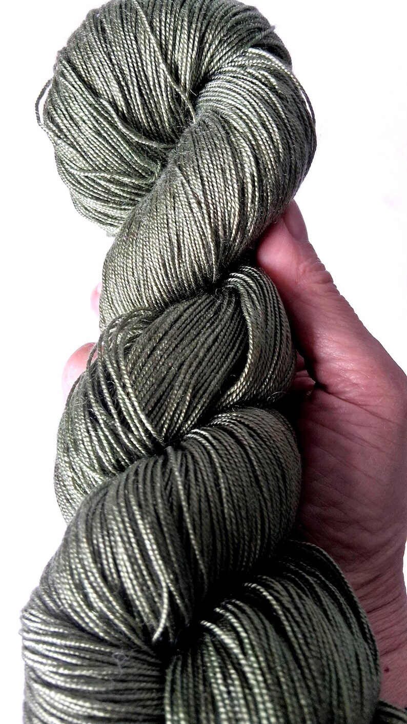May include: A hand holds a skein of olive green yarn. The yarn is tightly wound, revealing individual strands with a subtle sheen. The colour is a muted, earthy tone, ideal for various knitting or crochet projects.