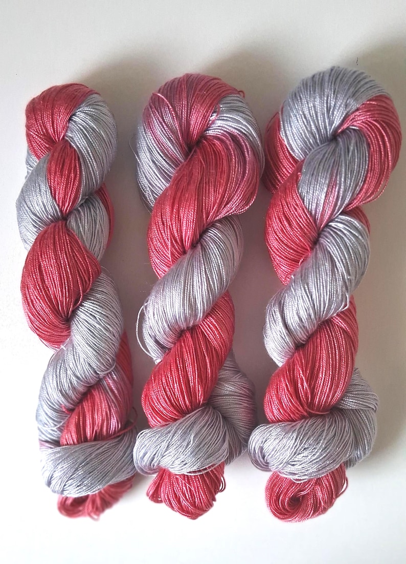 Puede incluir: Tres madejas de hilo en una configuraci&oacute;n retorcida. El hilo es una combinaci&oacute;n de plata y un vibrante color rojo-rosa. Las madejas est&aacute;n dispuestas una al lado de la otra sobre un fondo blanco, mostrando el color y la textura del hilo.