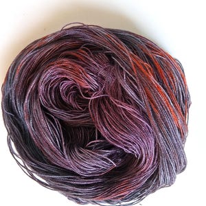 May include: A skein of hand-dyed yarn in shades of purple, gray, and orange. The yarn is tightly wound, showing the individual strands and color variations. The yarn is on a white background.