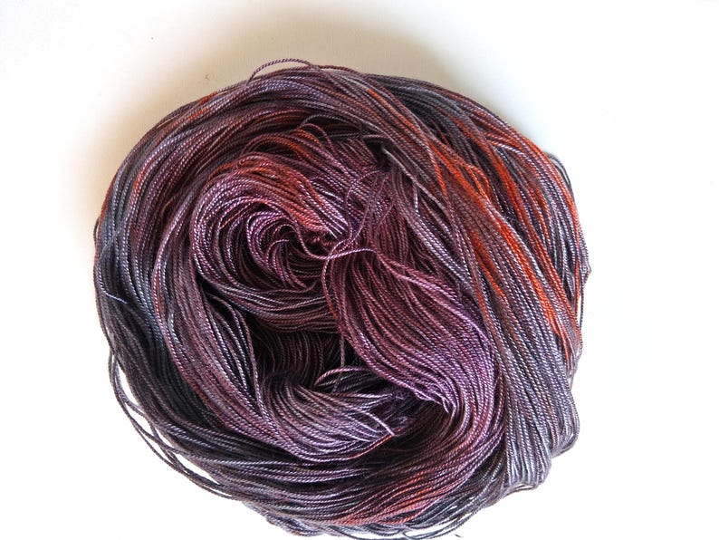 May include: A skein of hand-dyed yarn in shades of purple, gray, and orange. The yarn is tightly wound, showing the individual strands and color variations. The yarn is on a white background.