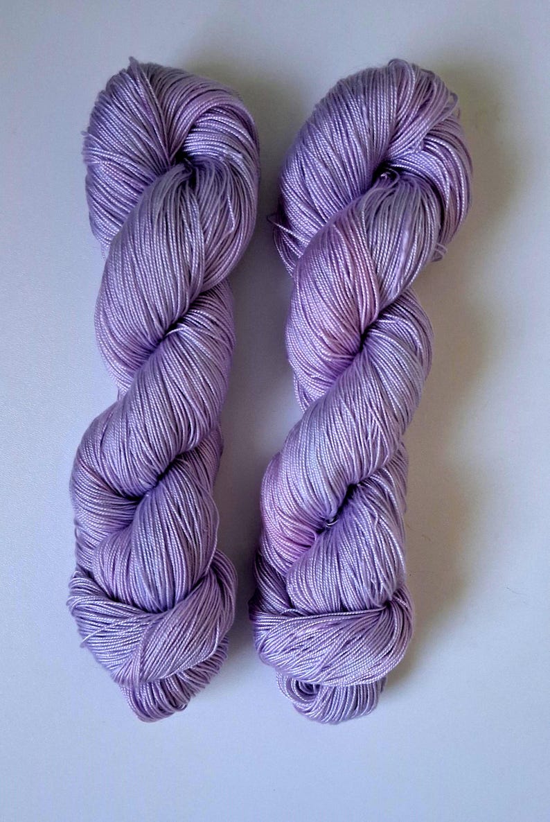 May include: Two skeins of lavender yarn are shown against a white backdrop. The yarn is tightly twisted, revealing individual strands. The colour is a soft, light purple with subtle tone variations, suggesting a high-quality fibre for knitting or crochet projects.