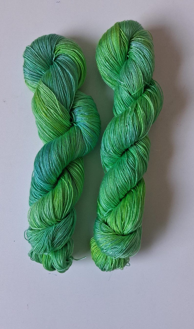 May include: Two skeins of green yarn with a gradient of colours from light to dark. The yarn is twisted into a rope-like shape, with a mix of light green, dark green, and a hint of blue.