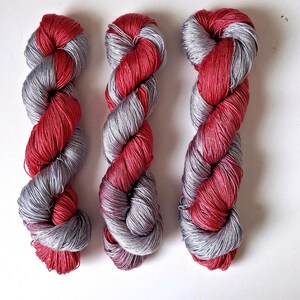 Puede incluir: Tres madejas de hilo con un dise&ntilde;o retorcido. El hilo combina colores rojo y gris plateado. Las madejas est&aacute;n dispuestas verticalmente sobre un fondo blanco. El hilo parece suave y adecuado para proyectos de tejido o ganchillo.