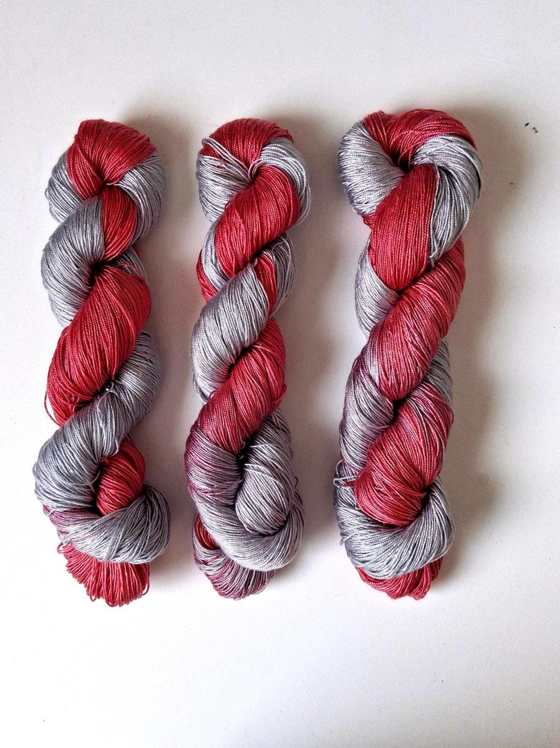 Puede incluir: Tres madejas de hilo con un dise&ntilde;o retorcido. El hilo combina colores rojo y gris plateado. Las madejas est&aacute;n dispuestas verticalmente sobre un fondo blanco. El hilo parece suave y adecuado para proyectos de tejido o ganchillo.