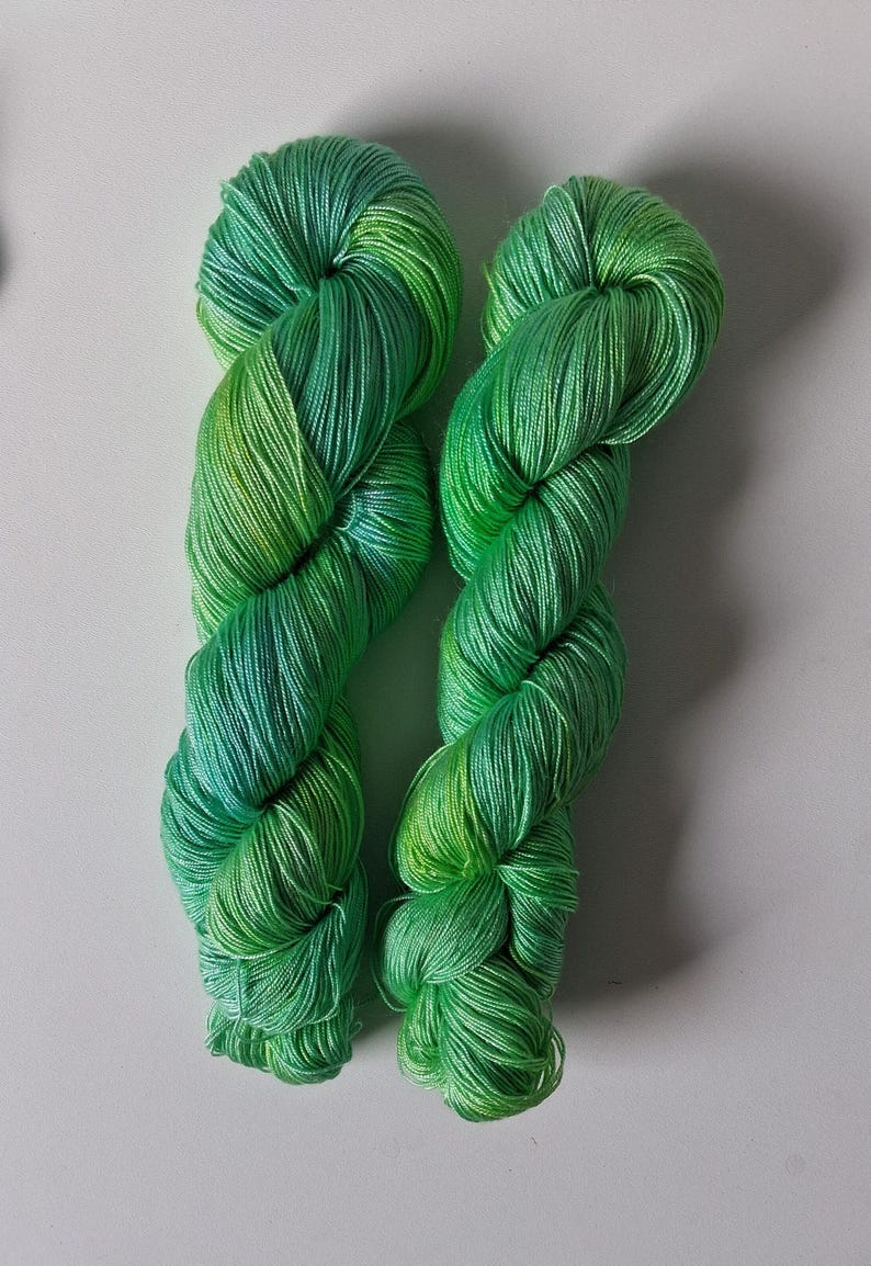 Peut inclure: Deux &eacute;cheveaux de fil vert vif avec un effet d&eacute;grad&eacute;, passant du vert jaune clair &agrave; un vert plus profond et plus riche. Le fil est &eacute;troitement enroul&eacute;, mettant en valeur sa texture et ses variations de couleur. Id&eacute;al pour les projets de tricot ou de crochet.