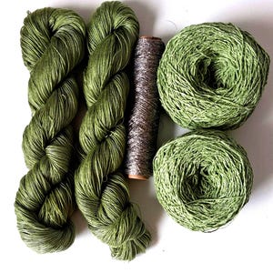May include: Two skeins of olive green yarn, two round balls of olive green yarn, and one central spool of metallic silver thread. The yarn has a smooth, lustrous texture, while the metallic thread features a shimmering, reflective finish.