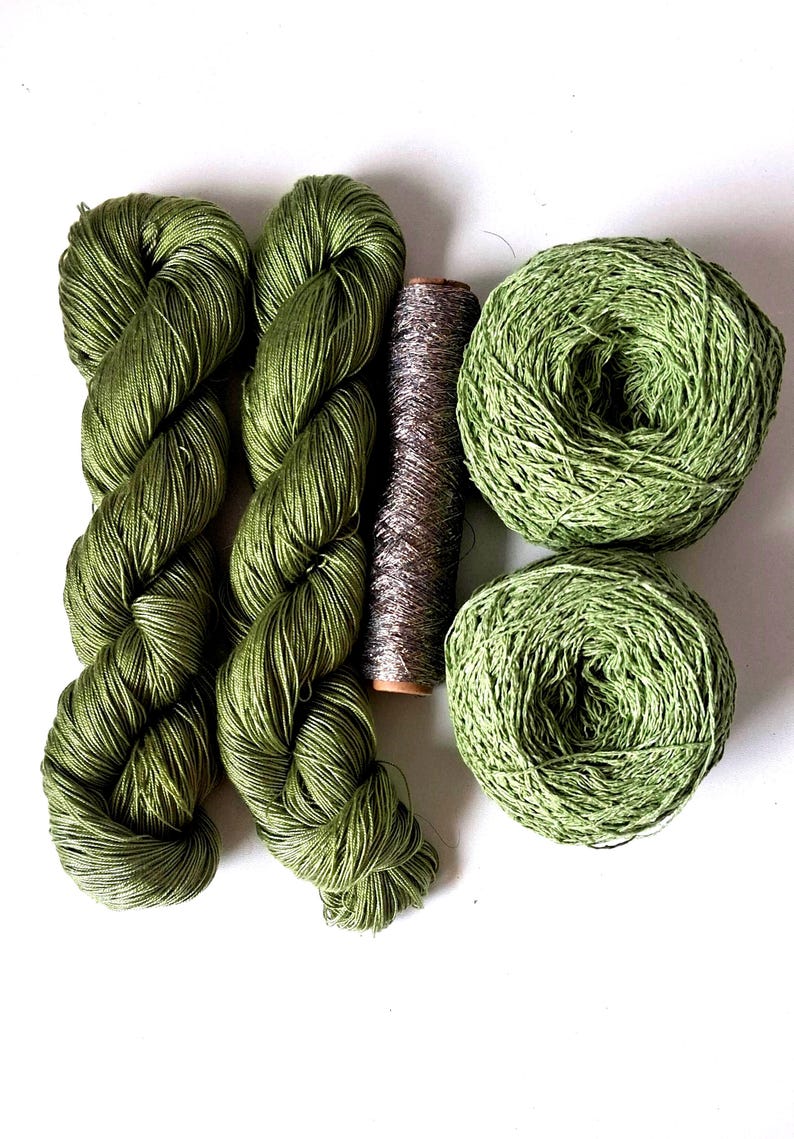 May include: Two skeins of olive green yarn, two round balls of olive green yarn, and one central spool of metallic silver thread. The yarn has a smooth, lustrous texture, while the metallic thread features a shimmering, reflective finish.