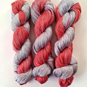 Puede incluir: Tres madejas de hilo en un patr&oacute;n retorcido, rojo y gris. Las madejas est&aacute;n dispuestas verticalmente sobre un fondo blanco. El hilo parece suave y adecuado para proyectos de tejido o ganchillo.