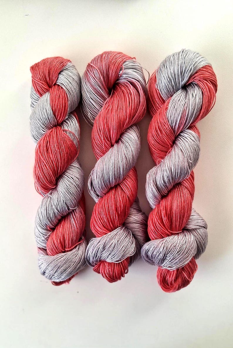 Puede incluir: Tres madejas de hilo en un patr&oacute;n retorcido, rojo y gris. Las madejas est&aacute;n dispuestas verticalmente sobre un fondo blanco. El hilo parece suave y adecuado para proyectos de tejido o ganchillo.