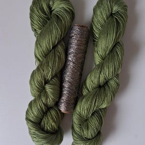 May include: Two skeins of olive green yarn with a smooth, lustrous finish are positioned vertically, flanking a single cylindrical spool of metallic silver thread that features a textured, shimmering appearance between the two green yarn bundles.