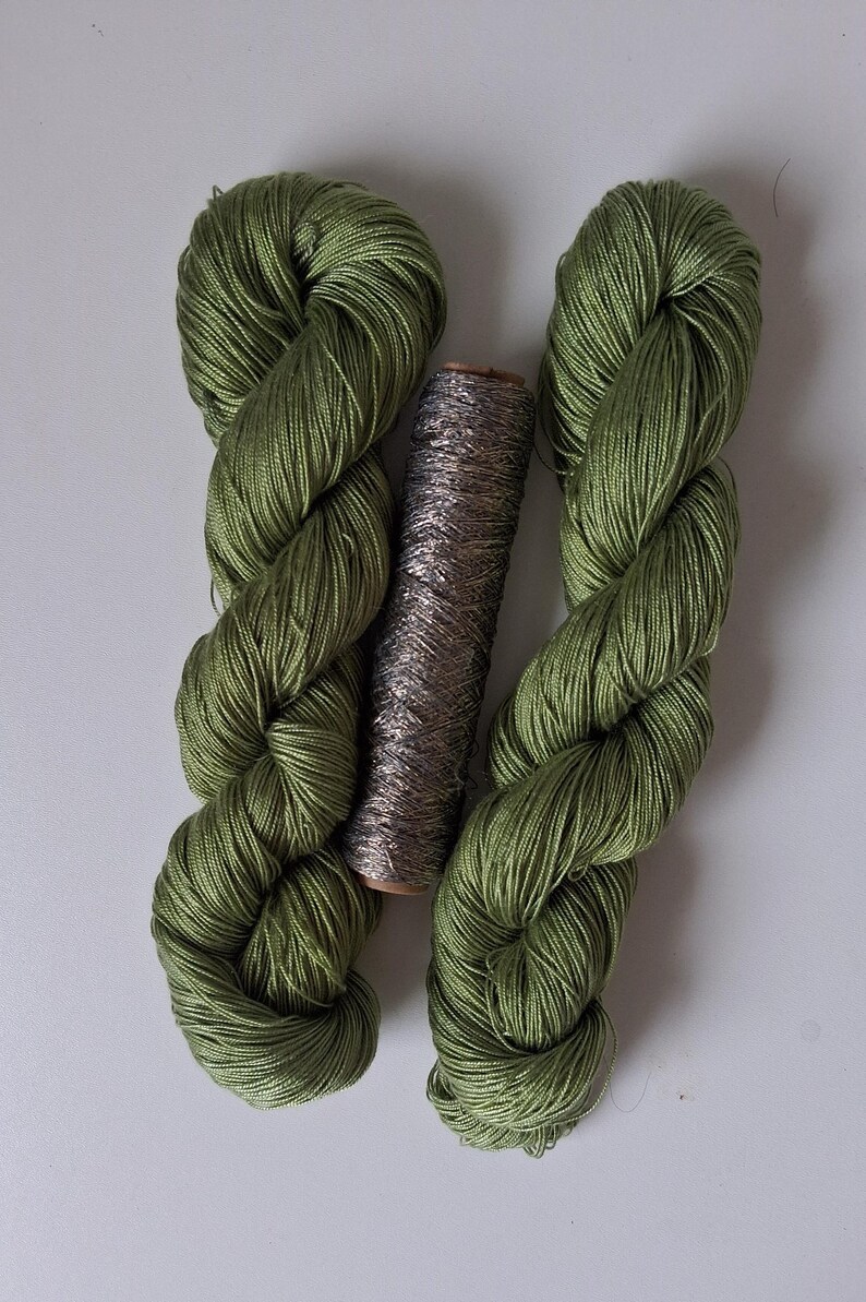 May include: Two skeins of olive green yarn with a smooth, lustrous finish are positioned vertically, flanking a single cylindrical spool of metallic silver thread that features a textured, shimmering appearance between the two green yarn bundles.