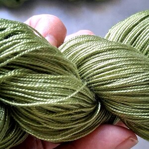 May include: Close-up of two skeins of green yarn. The yarn is a light, olive green color and appears to be made of a soft, silky material. The yarn is tightly wound and has a slight sheen. Ideal for knitting and crochet projects.