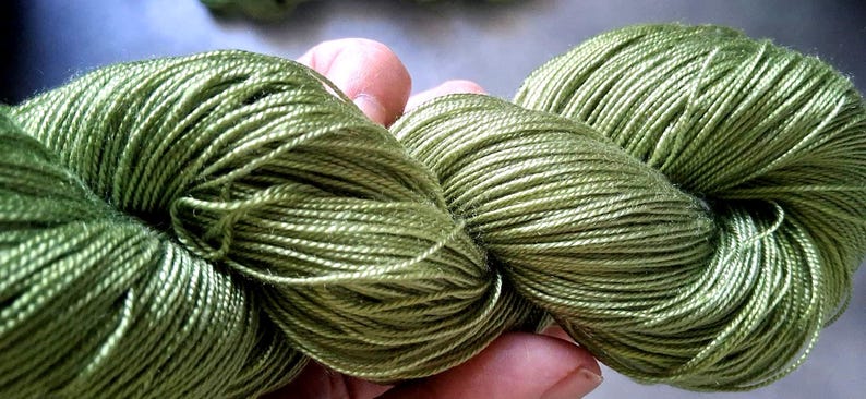 May include: Close-up of two skeins of green yarn. The yarn is a light, olive green color and appears to be made of a soft, silky material. The yarn is tightly wound and has a slight sheen. Ideal for knitting and crochet projects.